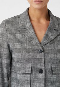 Blazer gris avec un motif à carreaux texturé, col à revers, deux grandes poches et boutons noirs. Le tissu semble net et structuré.