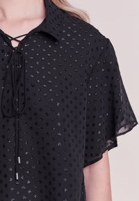 Blouse noire avec col, ornée de petits points noirs brillants. Dotée d'un décolleté lacé et de manches légères. Tissu léger.