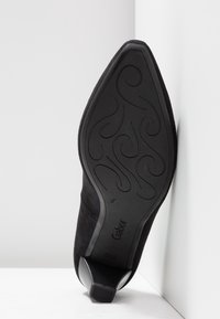 Chaussure à talon haut en daim noir avec un bout pointu et une semelle texturée, dotée d'un petit talon carré et d'accents latéraux incurvés.