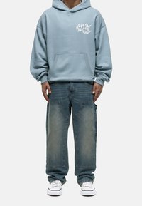 Sudadera con capucha azul claro con bolsillo frontal y texto gráfico. Vaqueros de corte holgado en azul deslavado con detalles únicos en los bolsillos. Zapatillas blancas.