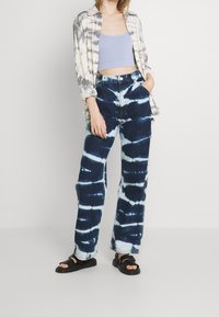 Denimbyxor med ett blått och vitt tie-dye-mönster, högmidjade, avslappnad passform, kombinerade med en ljusgrå crop top och en rutig skjorta.