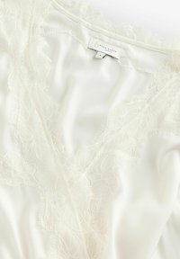 Blouse en satin crème avec bordure en dentelle, présentant un design à col en V et une texture douce et lisse. Étiquette visible avec les informations de marque et de taille.