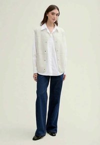 Gilet bianco imbottito con bottoni a pressione, indossato sopra una camicia bianca con colletto. Abbinato a jeans a gamba larga blu scuro e scarpe nere. Design semplice.