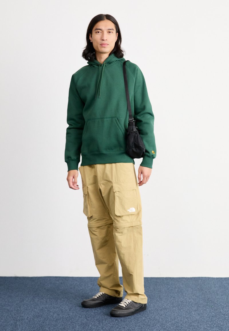 Sudadera verde con capucha hecha de algodón, combinada con pantalones cargo color caqui que cuentan con múltiples bolsillos. Acentuada con un bolso de hombro negro y zapatillas oscuras.