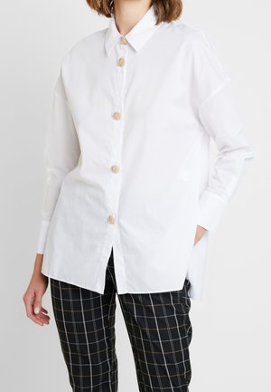 Femme portant une chemise blanche oversize à boutons dorés texturés et un pantalon à carreaux noir, main dans la poche, sur un fond uni.