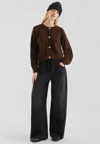 Bruine gebreide cardigan met een ruitpatroon, voorzien van gouden knopen, gecombineerd met brede zwarte broek en een zwarte beanie.