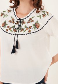 Blouse blanche avec broderie florale en orange et vert, présentant un col rond, une bordure noire et des franges noires au niveau du col.