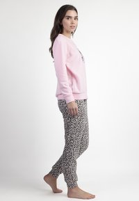 Mujer con una blusa de manga larga rosa y pantalones de leopardo, de pie descalza, mirando hacia la derecha contra un fondo blanco liso.