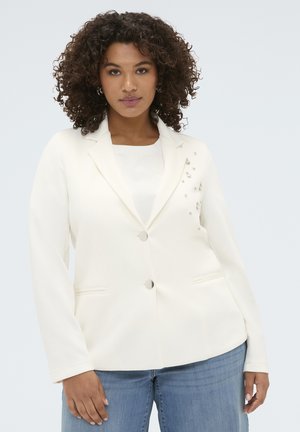 Vrouw met krullend haar die een witte blazer met zilveren versieringen en een blauwe spijkerbroek draagt, kijkt naar voren tegen een effen achtergrond.