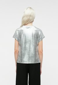 En blond person med langt hår står med ryggen til, iført en skinnende sølvfarvet kortærmet top og løse sorte bukser foran en ensfarvet baggrund.