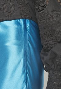 Haut noir à manches longues avec des détails en dentelle complexes, associé à une jupe en satin bleu brillant avec une fermeture éclair latérale visible.