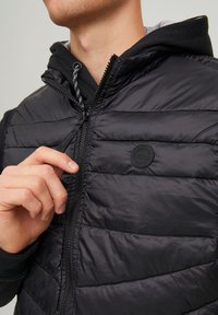 Gilet imbottito nero con chiusura a zip, dotato di una patch con logo e un colletto alto. Il rivestimento interno presenta un cappuccio grigio visibile sotto.
