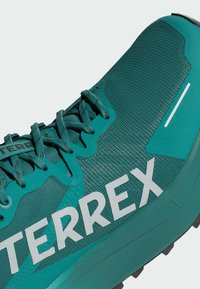 Scarpa sportiva turchese con una parte superiore testurizzata, caratterizzata da un prominente logo bianco "TERREX" e un materiale in mesh traspirante. Include lacci e un accentuazione riflettente.