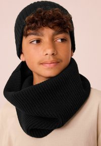 Sort strikket hue og bred infinity-scarf, begge med ribbet struktur. Scarf'en hænger løst rundt om halsen. Modellen bærer en lyse farvet skjorte.