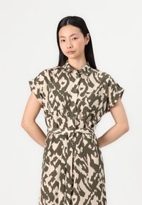 Abito camicia fino al ginocchio in beige con stampa animale verde oliva. Caratterizzato da maniche corte, chiusura con bottoni e cintura legata in vita.