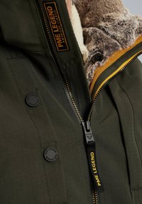 Groene jas met een met bont gevoerde kraag, voorzien van een zwarte rits, oranje accenten en logo knopen. Gelaagd stof met subtiele strepen.