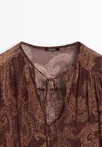 Blusa marrone con motivo paisley e tessuto testurizzato, caratterizzata da un collo a V e chiusura con lacci sul davanti; maniche lunghe e arricciate.