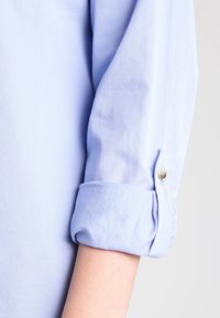 Ljust blå skjorttyg med uppvikt ärm och en knappdetalj. Slät struktur; enkel design med rena linjer och utan mönster.