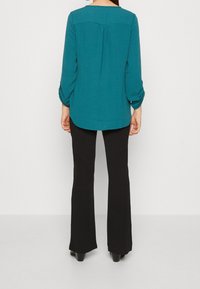 Blusa teal a maniche lunghe con scollatura rotonda, dettaglio a pieghe sul retro e polsini arrotolati. Abbinata a pantaloni neri a campana.