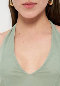 Γυναίκα που φοράει ανοιχτό πράσινο τοπ με τιράντες στο λαιμό και μεγάλα χρυσά σκουλαρίκια κρίκους, εμφανίζοντας λαιμό, κλείδα και χείλη, με ορατά σπίλους δέρματος στο στήθος.