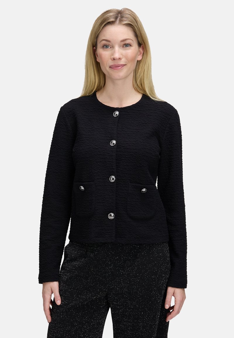 Blondynka uśmiechająca się, ubrana w strukturalną czarną marynarkę z guzikami i kieszeniami z przodu oraz błyszczące czarne spodnie, stojąca na białym tle.