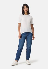 Witte T-shirt met korte mouwen, gemaakt van een lichte stof, gecombineerd met blauwe denim jeans. Model staat met witte sneakers op een effen achtergrond.