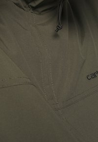 Grüne, leichte Oberbekleidung besteht aus strukturiertem Stoff, hat einen hohen Kragen und subtile Nähte. Der Markenname wird in dunkler Schrift angezeigt.