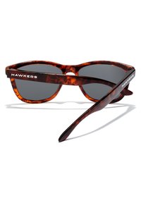 Ochelari de soare cu rame de țestoasă și lentile întunecate, având un cadru curbat și brațe elegante. Numele brandului "HAWKERS" este vizibil pe laterale.