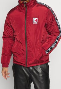 Red padded jacket with stand-up collar, zip closure, logo patch on chest, and contrasting black side stripes. Smooth, glossy texture.