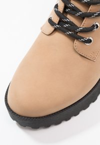 Botín de tobillo de imitación de cuero beige con punta redonda, suela de goma texturizada negra y cordones con patrón en blanco y negro.