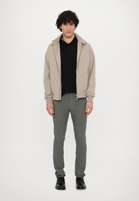Jachetă crem deschis cu fermoar peste un tricou polo negru, purtată cu pantaloni oliu slim-fit și pantofi negri, stând pe un fundal alb.