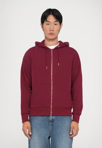 Haut à capuche zippé bordeaux en tissu doux, avec une capuche à cordon de serrage et des poches avant. Porté avec un jean bleu clair.