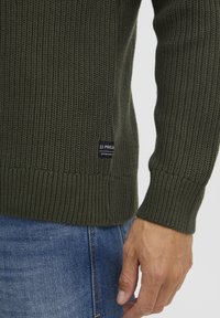 Dunkelgrüner, gestrickter Pullover mit Rippbündchen und -saum. Besitzt ein kleines schwarzes Etikett mit weißem Text auf der linken Seite. Die Textur wirkt dick und strukturiert.