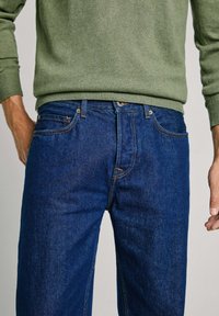 Jeans de denim azul oscuro con un corte relajado, diseño de cinco bolsillos y costura en naranja contrastante, combinados con un suéter verde.