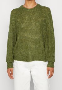 Grön stickad tröja med en strukturerad design, långa ärmar och rundad halsringning, kombinerad med vita byxor.