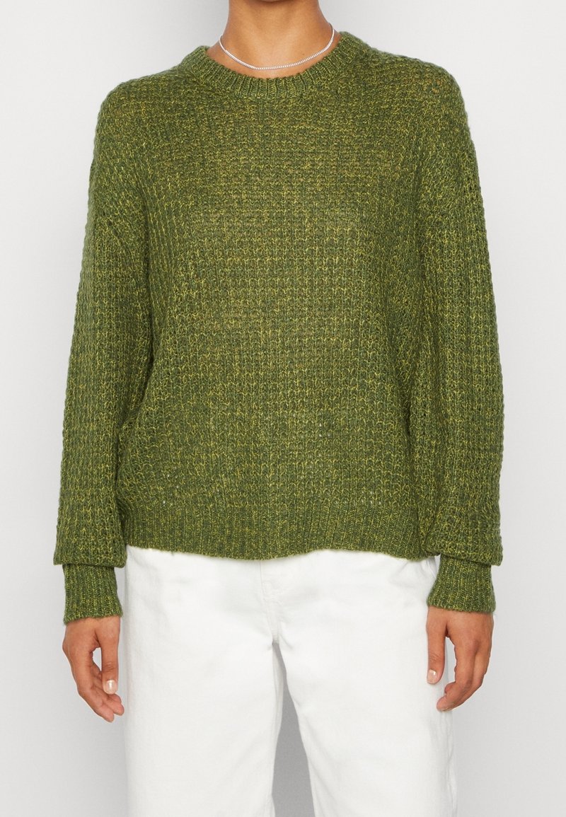 Grön stickad tröja med en strukturerad design, långa ärmar och rundad halsringning, kombinerad med vita byxor.