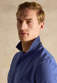 Zila polo krekls ar teksturētu apkakli un mazu melnu logotipu uz krūtīm. Iekļauj pogu rindu un īsām piedurknēm.