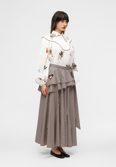 Γυναίκα που φοράει λευκό κεντητό πουκάμισο με φουσκωτά μανίκια και μακριά ριγέ φούστα με στρωμένο πεπλούμ, σε συνδυασμό με μαύρα φλατ παπούτσια, όρθια.