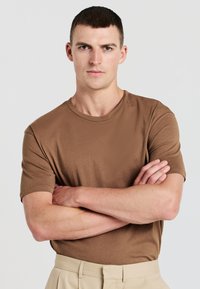 Giovane uomo con capelli corti e scuri, vestito con una maglietta marrone e pantaloni beige, si trova con le braccia incrociate su uno sfondo neutro.