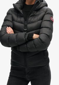 Persona che indossa un giubbotto invernale nero con chiusura a zip e una patch rossa sul manica, con le braccia incrociate, sopra un maglione nero e jeans scuri.