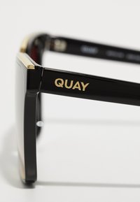 Črne sončne očala z debelimi pravokotnimi okvirji. Zlati poudarki blizu zaponke in vtisnjen logotip "QUAY" na strani. Polirana površina, temna stekla.