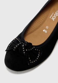 Schwarze Wildleder-Flatschuh mit runder Zehenpartie, einem Schleifenakzent, der mit kleinen Strasssteinen verziert ist, und einer gepolsterten Innensohle mit Perforationen.