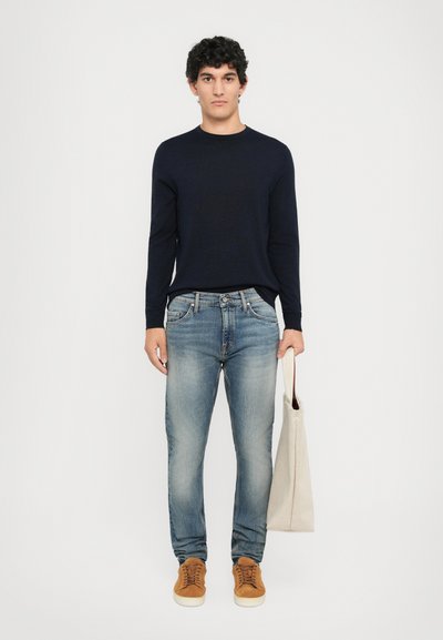 Young man standing, wearing a dark navy sweater, faded blue jeans, brown sneakers, holding a beige tote bag in his right hand.