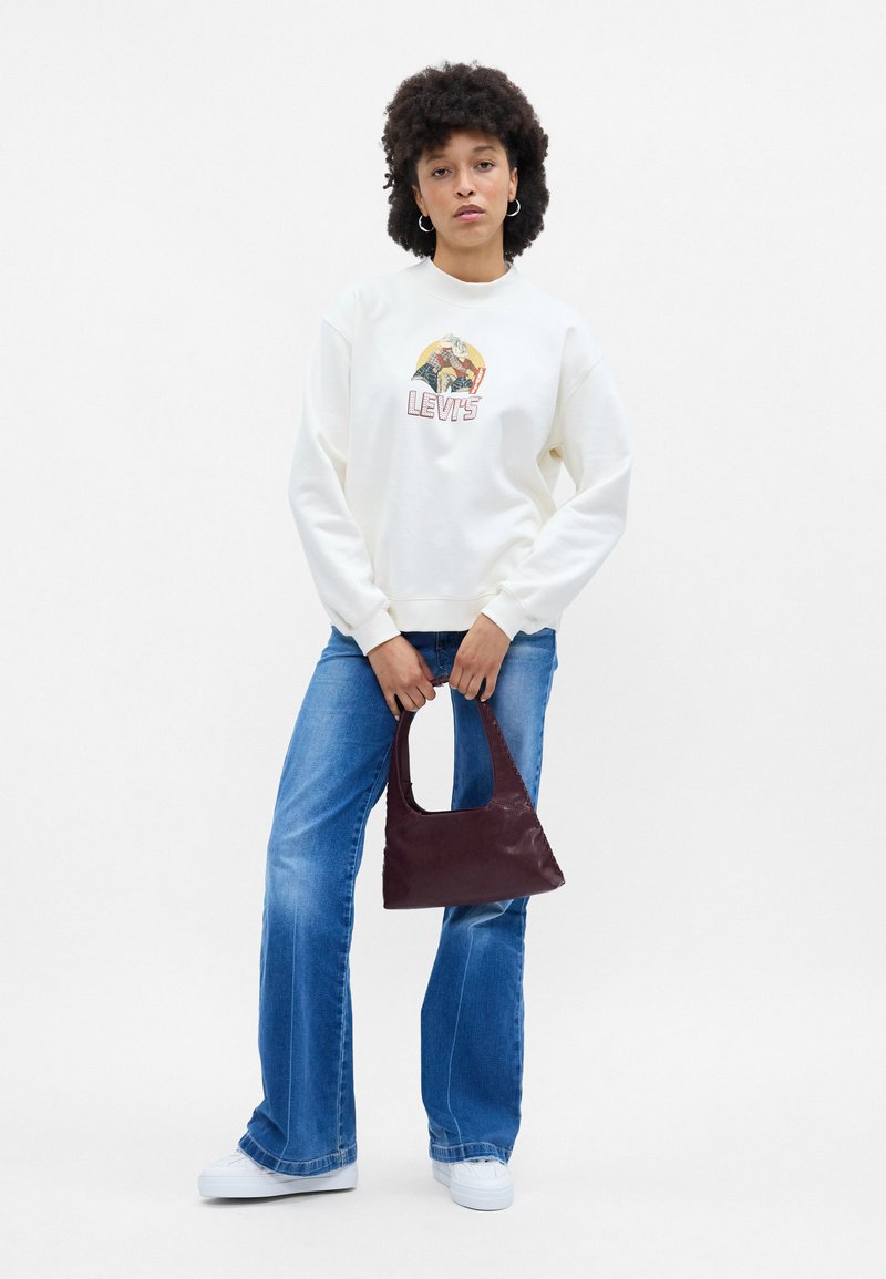 Sudadera de crema con estampado gráfico, jeans azules con piernas acampanadas y un pequeño bolso burdeos con un asa redondeada. Zapatillas blancas.