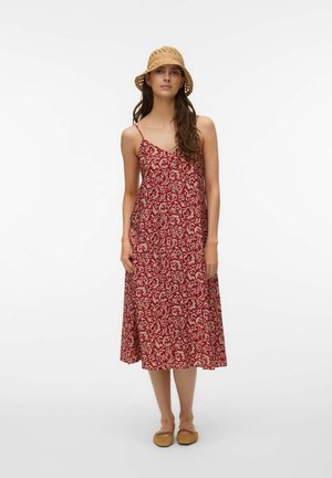 Femme portant une robe midi à fleurs rouge avec bretelles spaghetti, un chapeau beige tressé et des ballerines marron clair, debout devant un fond blanc.