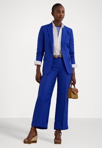 Un pantalon en lin bleu vif avec des jambes larges, une chemise à col blanc, un top rayé en dessous, et un sac en osier. Des sandales compensées marron complètent le look.