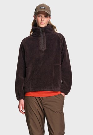 Jeune personne portant un pull polaire marron, une chemise orange en dessous, un pantalon marron et une casquette marron avec un logo, debout devant un fond uni.