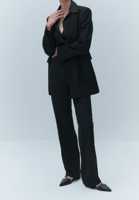 Black tailored blazer with structured shoulders, worn over a black blouse, paired with fitted black trousers and pointed black flats.