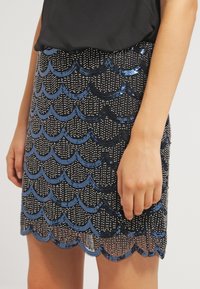 Mini-jupe noire avec motifs coquilles en sequins bleus et détails en perles argentées, portée avec un haut noir.