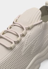 Gros plan sur une basket tricotée beige clair avec une tige épaisse semblable à une chaussette, des lacets élastiques en boucle et une semelle texturée.